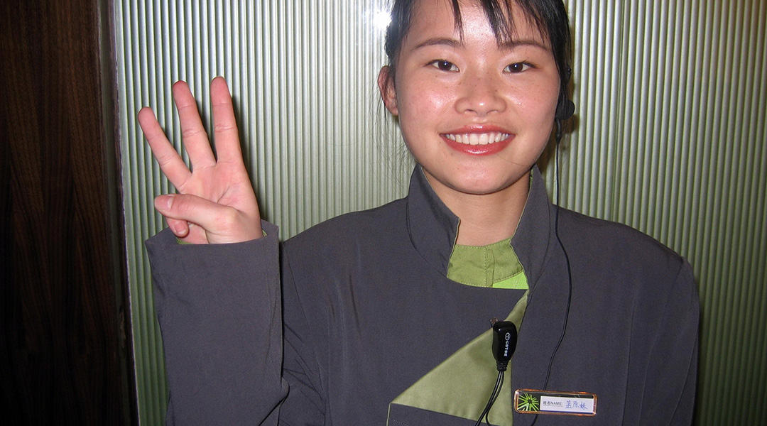 image of a woman dressed in dark purple with green accents giving a 3-finger symbol to the camera