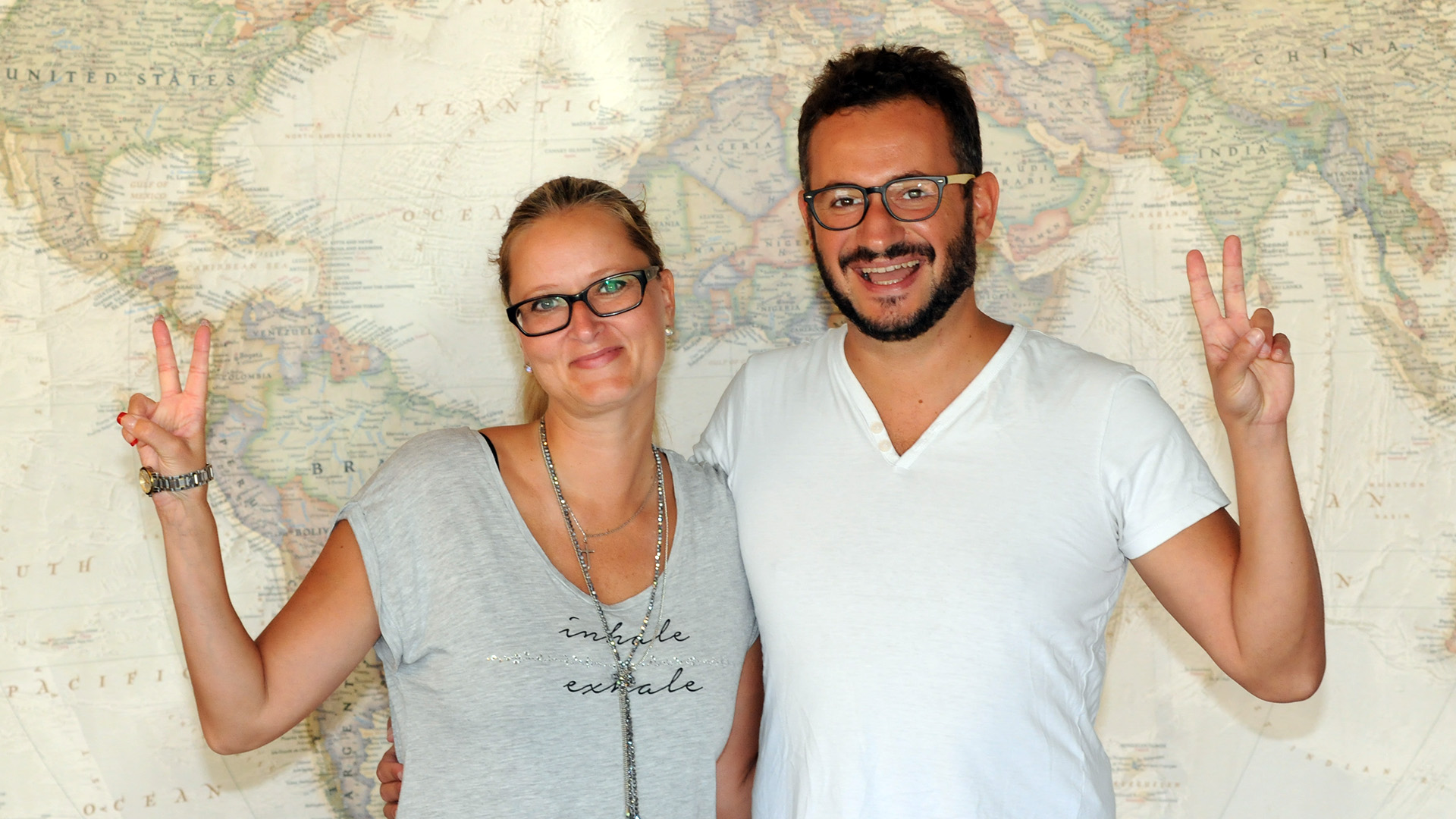 Two people standing in front of a ten foot wide map of the world, smiling and making peace signs