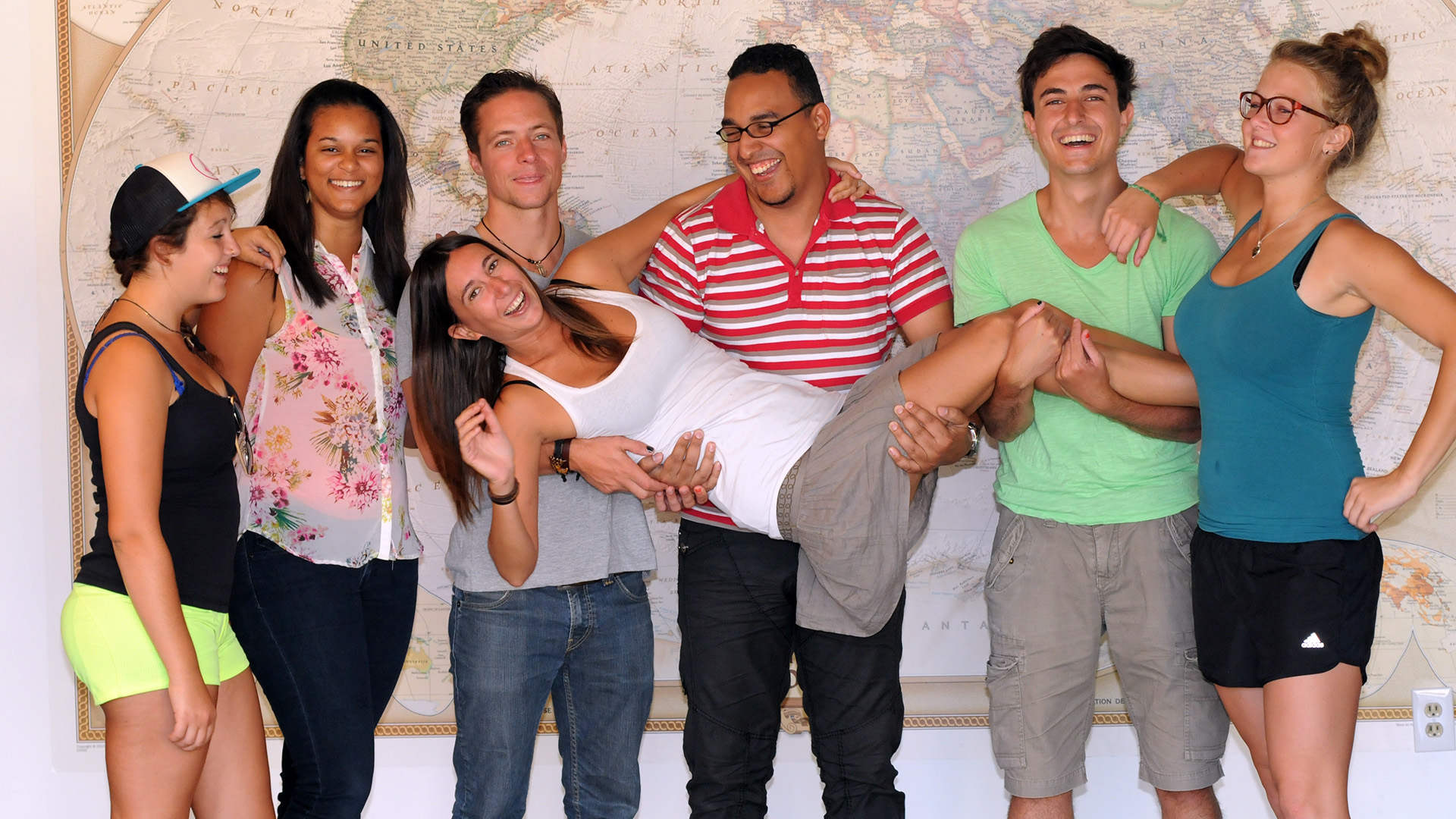 6 people standing in front of a world map, with a 7th person being carried by the group