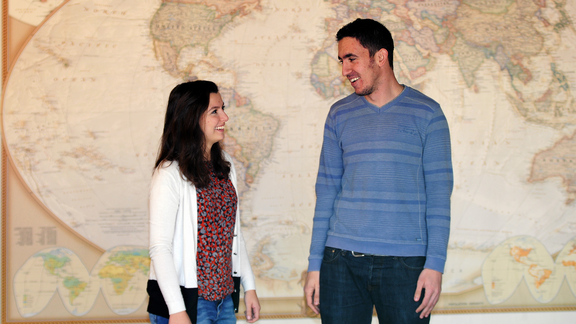 Rania and Moe stand in front of a world map and smile at each other