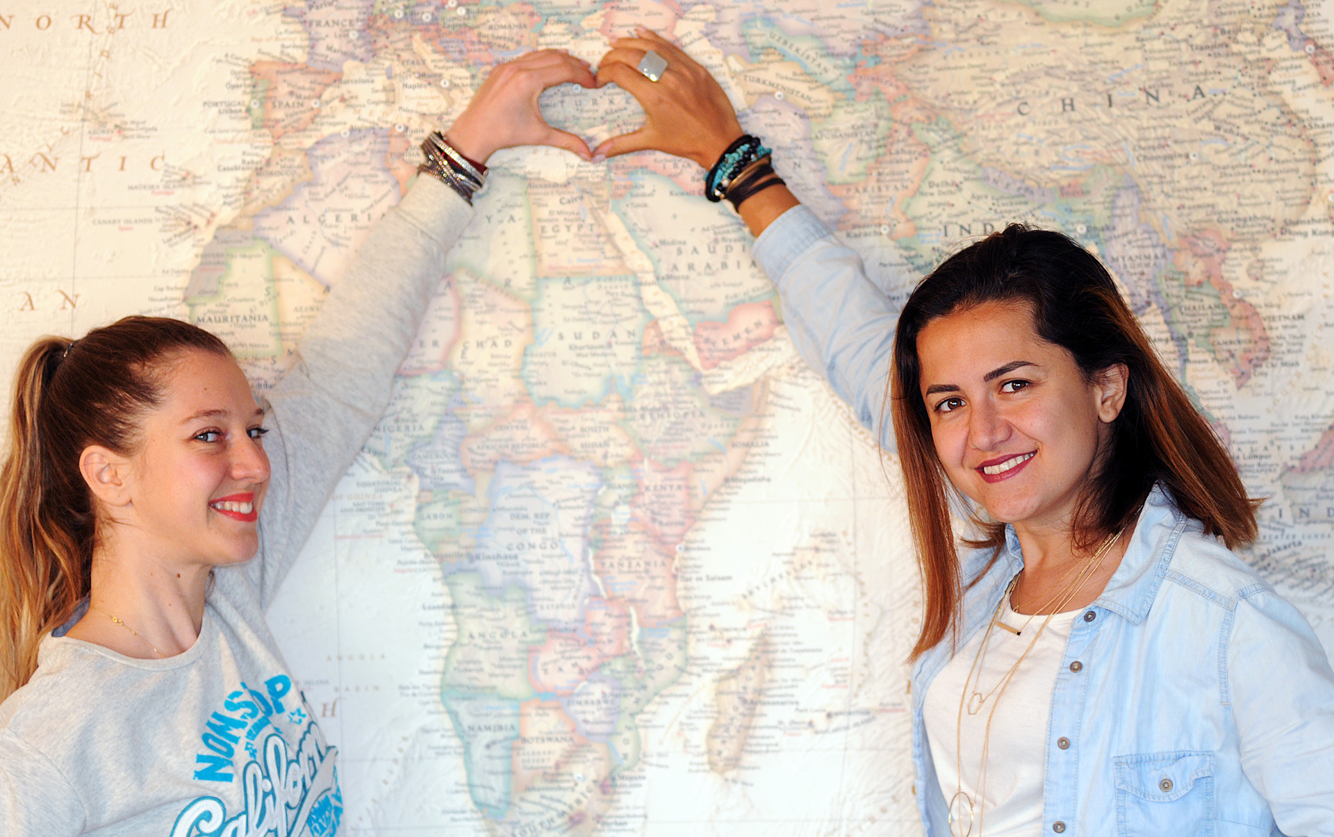 Pinar & Gizem in front of a large world map and making a heart shape around Turkey with their hands