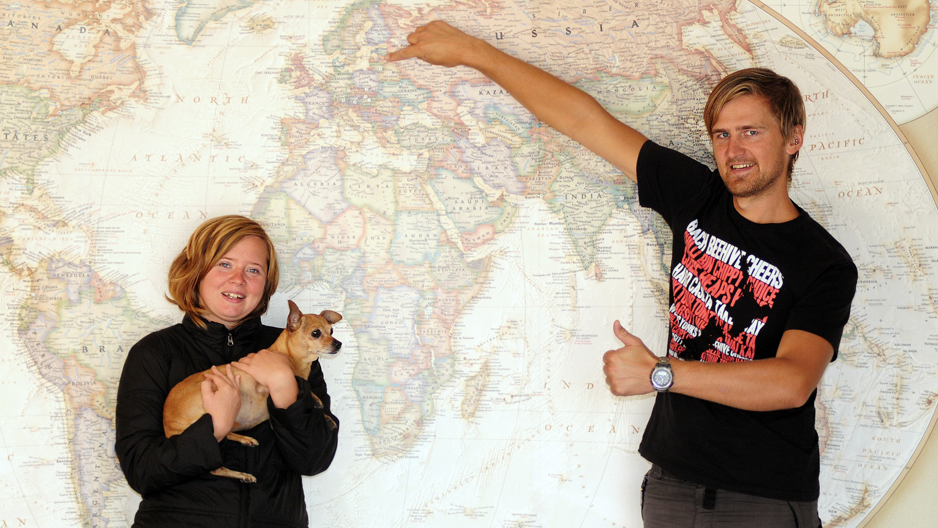 Vineta, Tiki & Krisjanis in front of a large world map. Vineta is holding Tiki and Krisjanis is pointing to Riga, Latvia on the world map