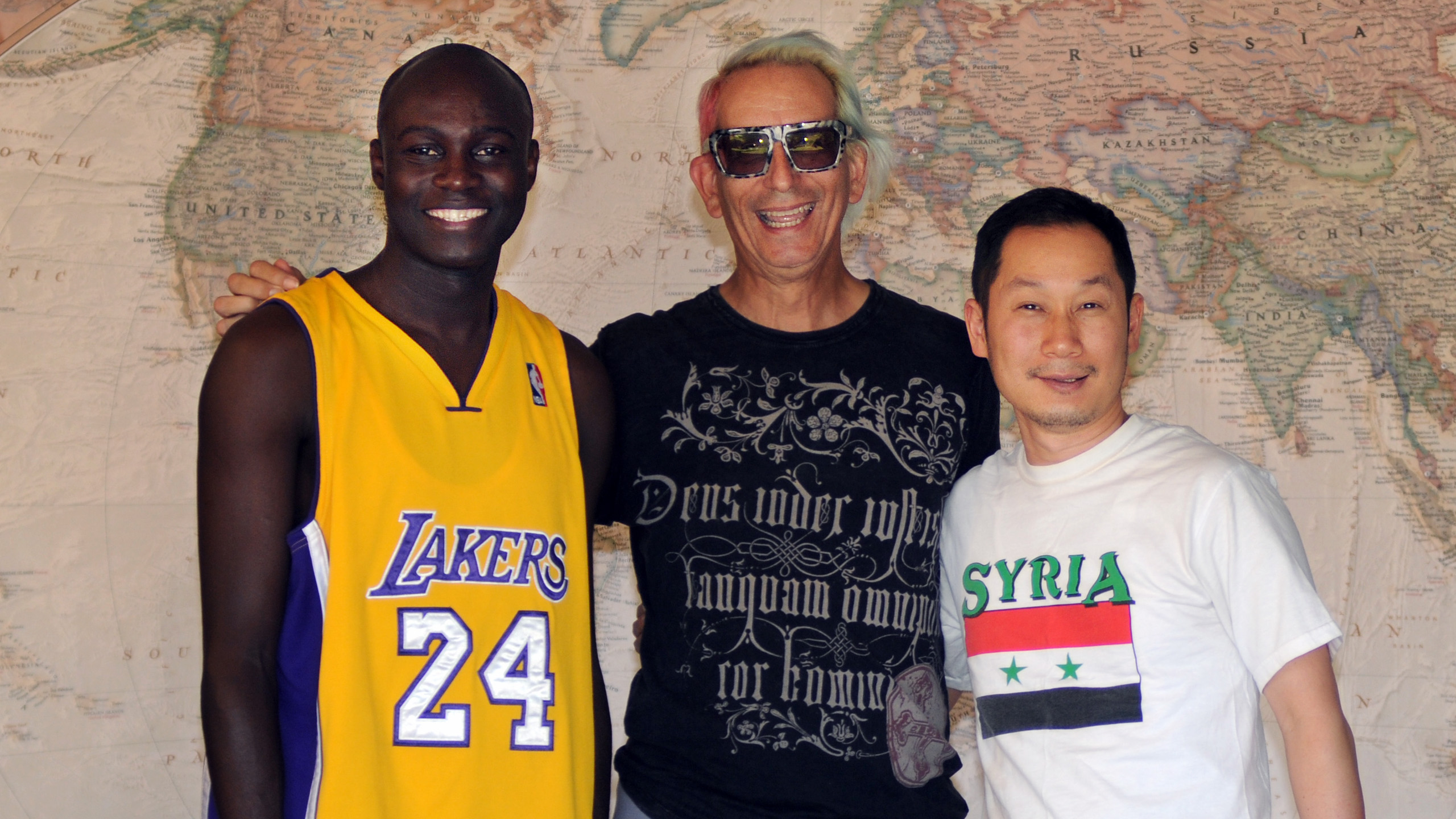 Abdoulaye Sane, Glenn Zucman & Terry Choi stand in front of a large world map. Abdoulaye wears a Laker jersey with #24, Kobe Bryant, Glenn wears a black t-shirt, and Terry wears a white t-shirt with the Syrian flag on it