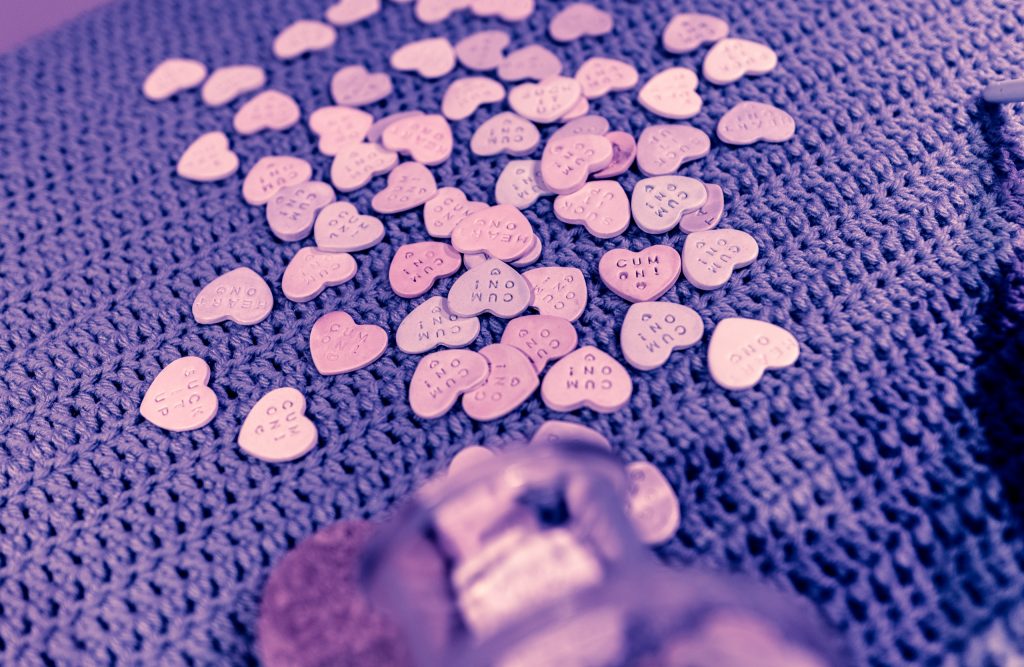 dozens of small ceramic candy hearts made by Claudia Solorzano on a crocheted table top including many repetitions of  "Cum On!" and "Suck it up"