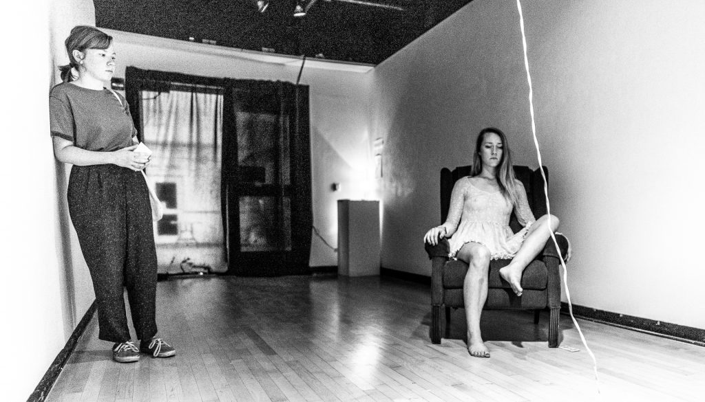 black-and-white photograph of Cortnee Brush sitting in a chair in the CSULB Merlino Gallery and a Gallery Visitor standing to the side and viewing Brush's performance work