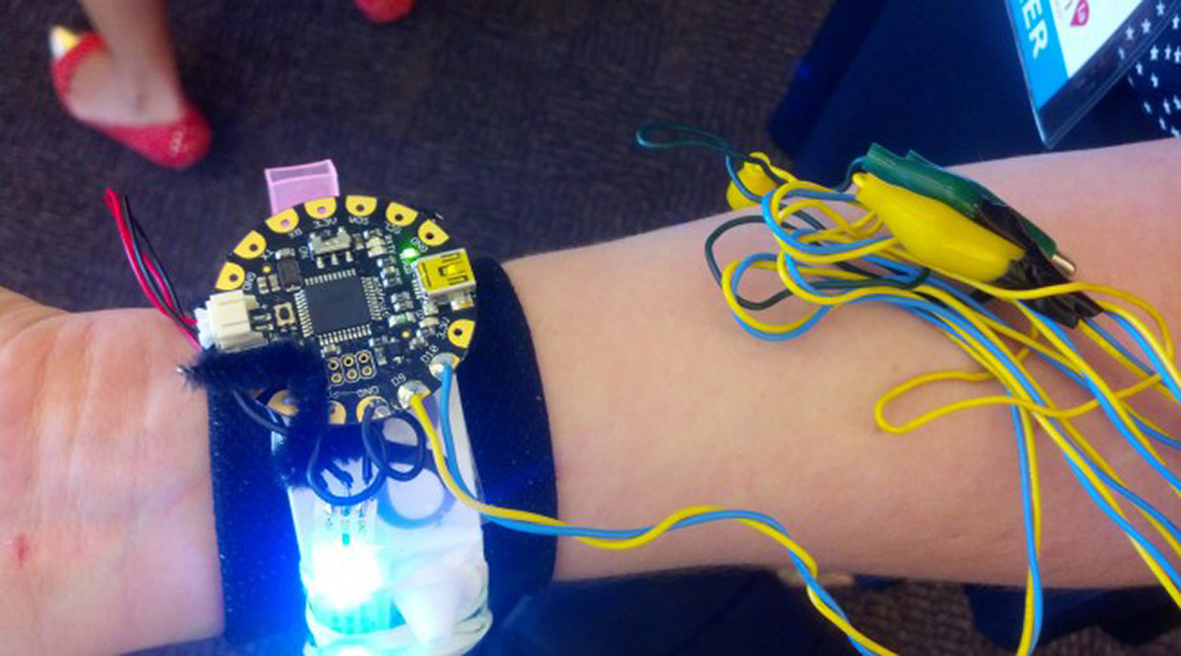 photo of a tiny circuit board on a wrist band lighting up a series of LEDs going around the wearer's wrist. Image is of a demo prototype and includes alligator clips connecting the electronics.