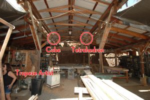 Photo of a workshop with a pole barn roof and various wood and metal machines. From the ceiling hang welded steel sculptures in the forms of articulated or "Disguised" Tetrahedron and Cube.