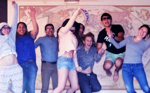 8 people jumping in the air and laughing in front of a large world map on New Year's Day 2016. One of the jumpers holds a "2016" sign in her hand