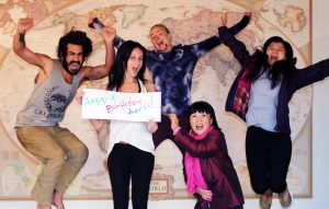 5 CouchSurfers jump in the air in front of a world map and holding a sign that reads "Happy Birthday Adriii - Runaway University"