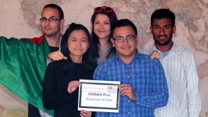 Mehdi, Yin Chien, Aylar, Jimmy & Sagar standing in front of a large world map and holding a sign that reads "Experience America, UGRAD Post, University of Utah" Mehdi is wearing the Algerian flag.