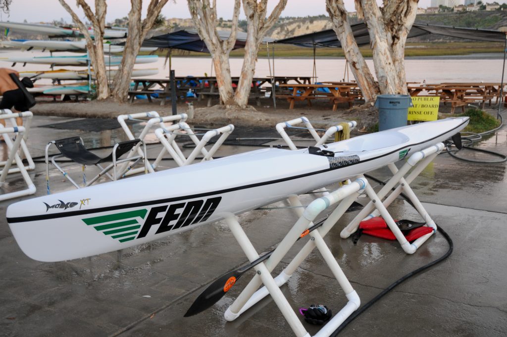 A Fenn surf ski or kayak sitting on slings on a concrete deck between a boat house and the beach.