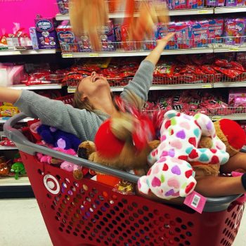 Marina Barnes in a red Target shopping cart and covered with plush animals, some of which she is tossing joyously into the air with her arms outstretched in celebration and her feet sticking out the end of the cart