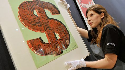 Image of an Andy Warhol dollar sign screen print in a frame being handled by a preparator in a black, Bohnam's London, t-shirt