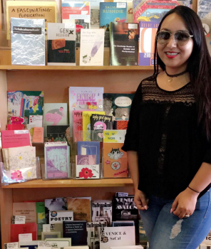 Janett Moctezuma standing in front of many zines and other small, local publications at Small World Books on the boardwalk at Venice Beach, CA