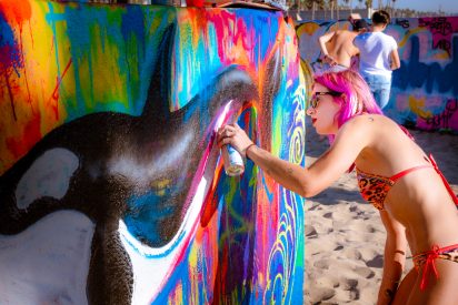 painter and street artist Lizzie Green spray painting a piece at the Venice Beach Art Walls. Green paints a whale in black and white, and surrounding the whale a brightly colored background field. Lizzie Green's own haircolor in fuschia and orange resonates with the colored spraypaint on the wall