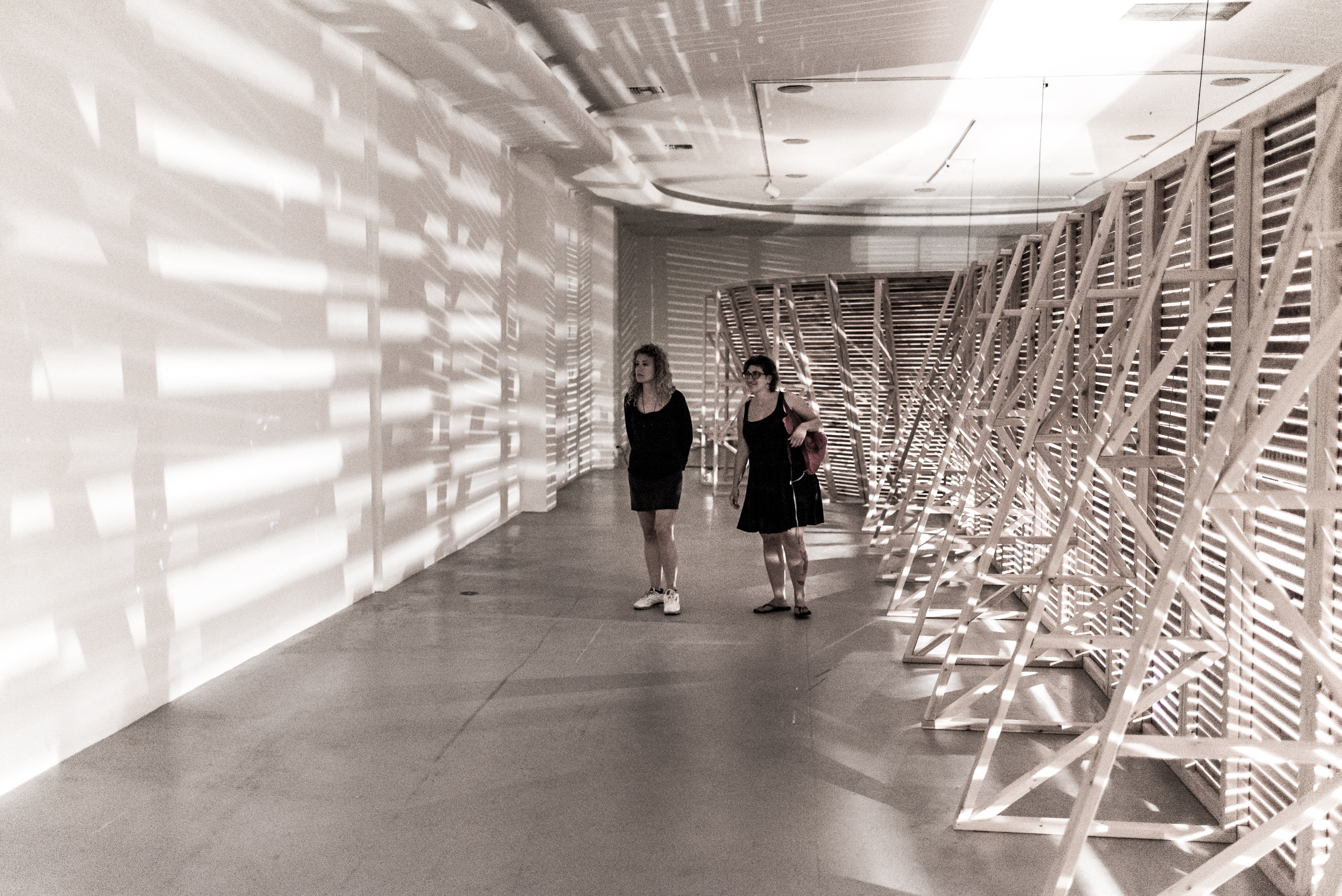 Jimena Sarno's installation "Home Away From" at Los Angeles Contemporary Exhibitions (LACE) on Hollywood Blvd in Los Angeles. The full-gallery installation consists of a lath-and-no-plaster wooden structure with a long corridor leading to a round room. The corridor and room have a few lights bulbs hanging like pendulums from long cords. This is the only light in the installation. Visitors can sway the lights like pendulums. The lights cast shadows inside the space and also through the lath they make geometric patters on the gallery walls on the other side. The piece is accompanied by an ambient music score that comes in through the lath.