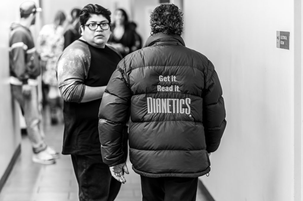 a man walks down the restroom corridor at Hollywood & Highland wearing a large, down windbreaker. On the back large embroidered letters read "Get it. Read it. Dianetics"
