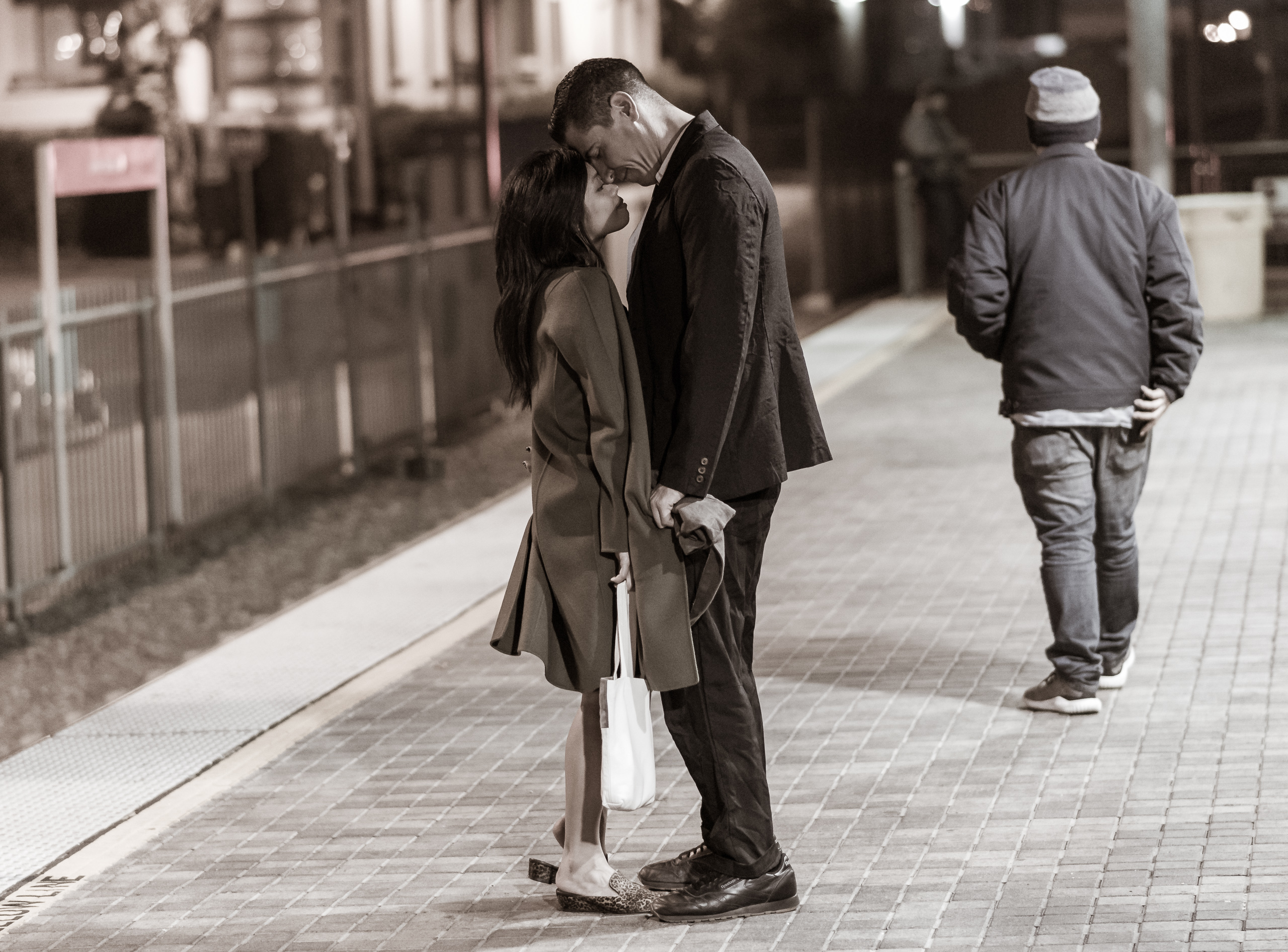 A man and a woman on the Gold Line platform at Union Station in Downtown Los Angeles. She wears an oversized coat which he holds the sleeves of as they press their heads to each other.