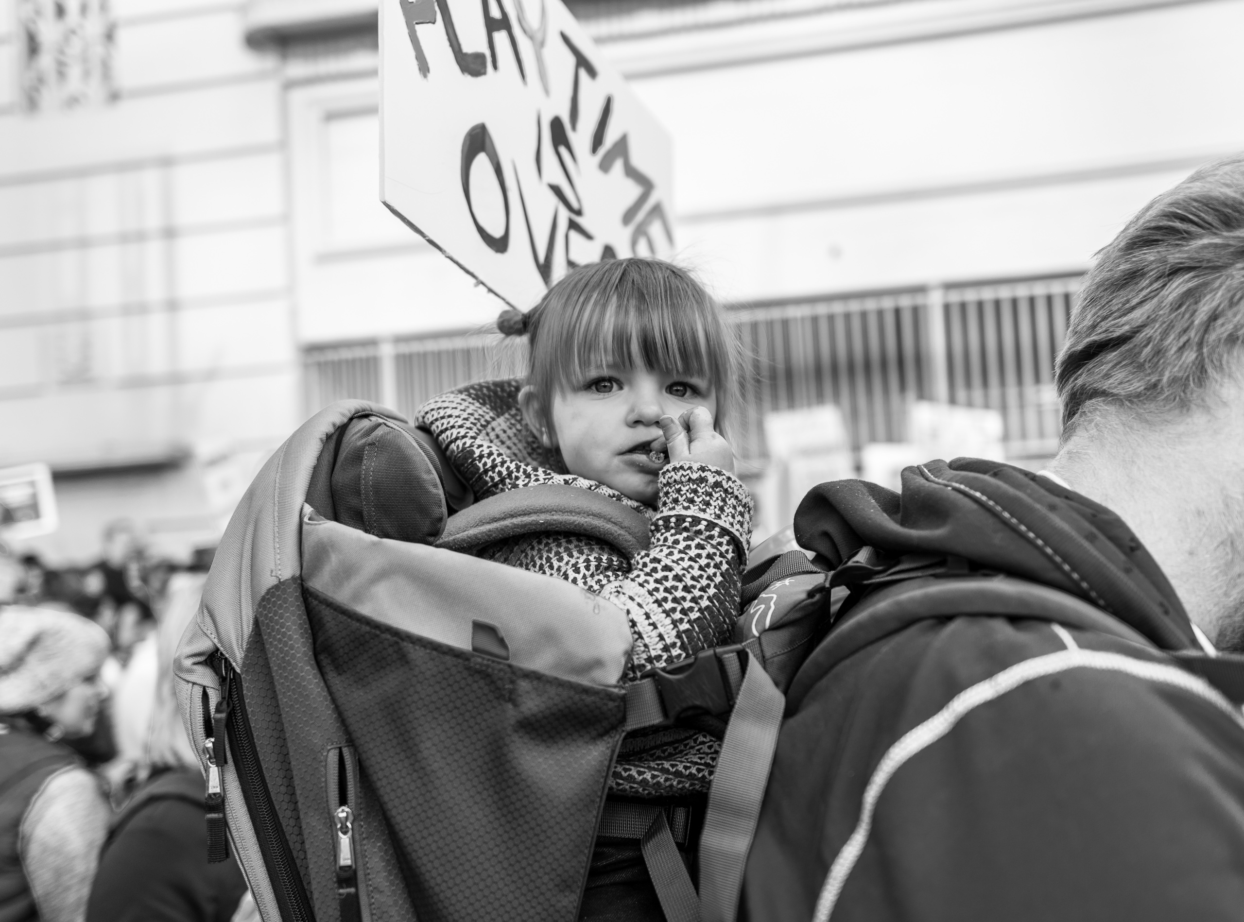 a young girl rides in a backpack on her dad's back and carries a sign that reads "playtime is over"