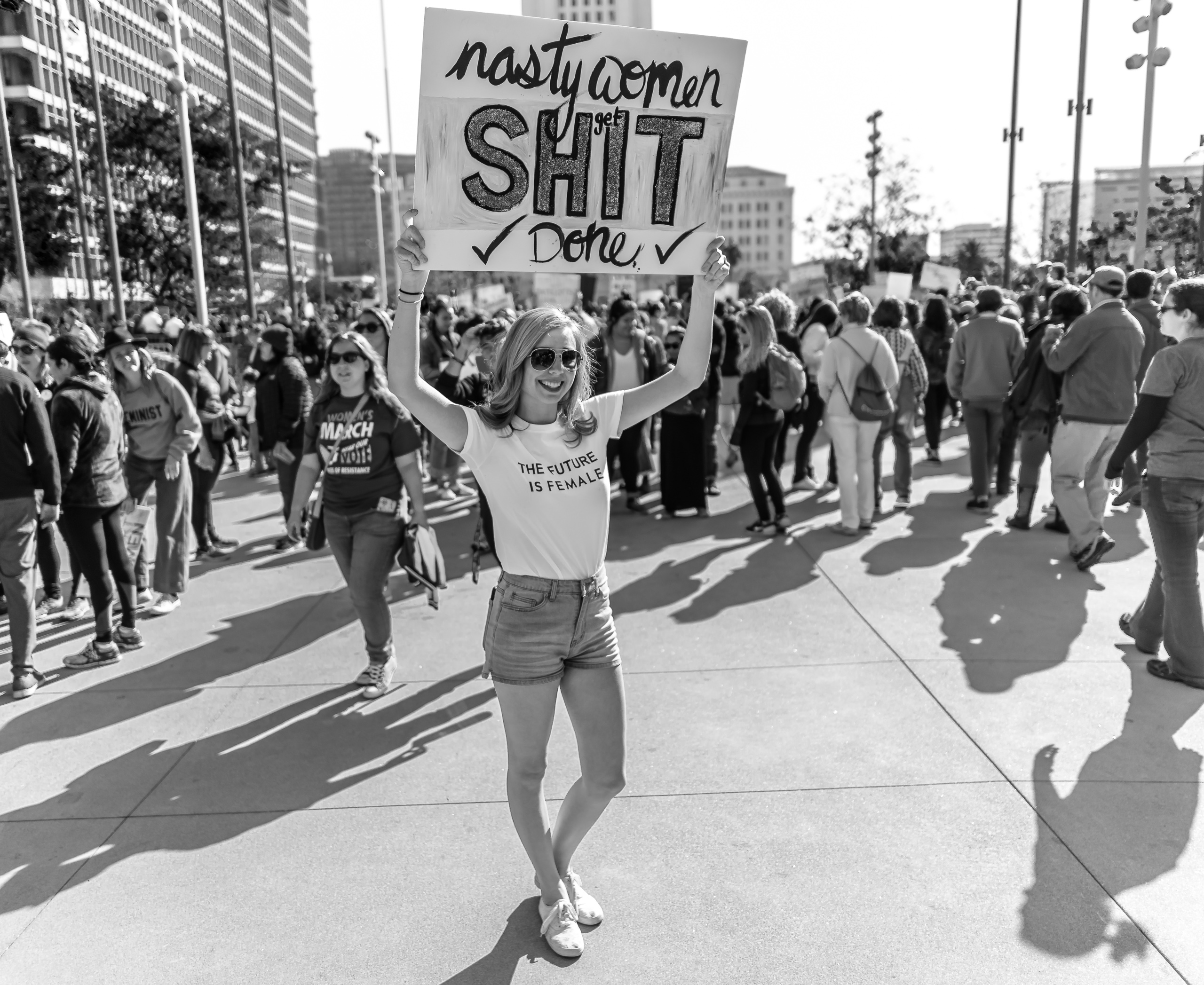 a woman standing in front of Los Angeles' City Hall and holding a sign in the air that reads "Nasty Women get shit done". Her t-shirt reads "the future is female"