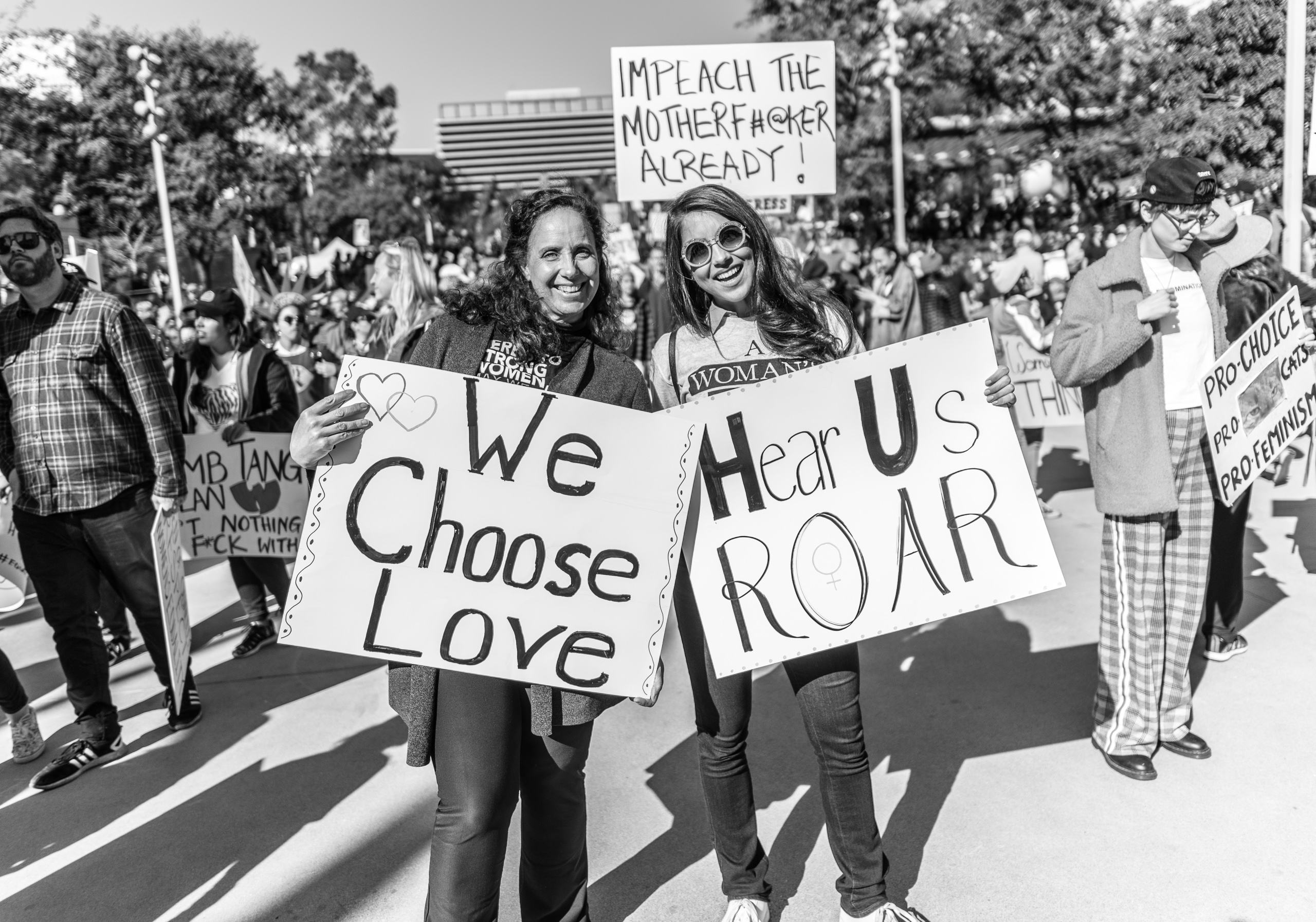 Two women standing together. One holds a sign that reads "We Choose Love". The other holds a sign that reads "Hear Us Roar".  Behind them another person holds up a sign reading "Impeach the motherfucker already"
