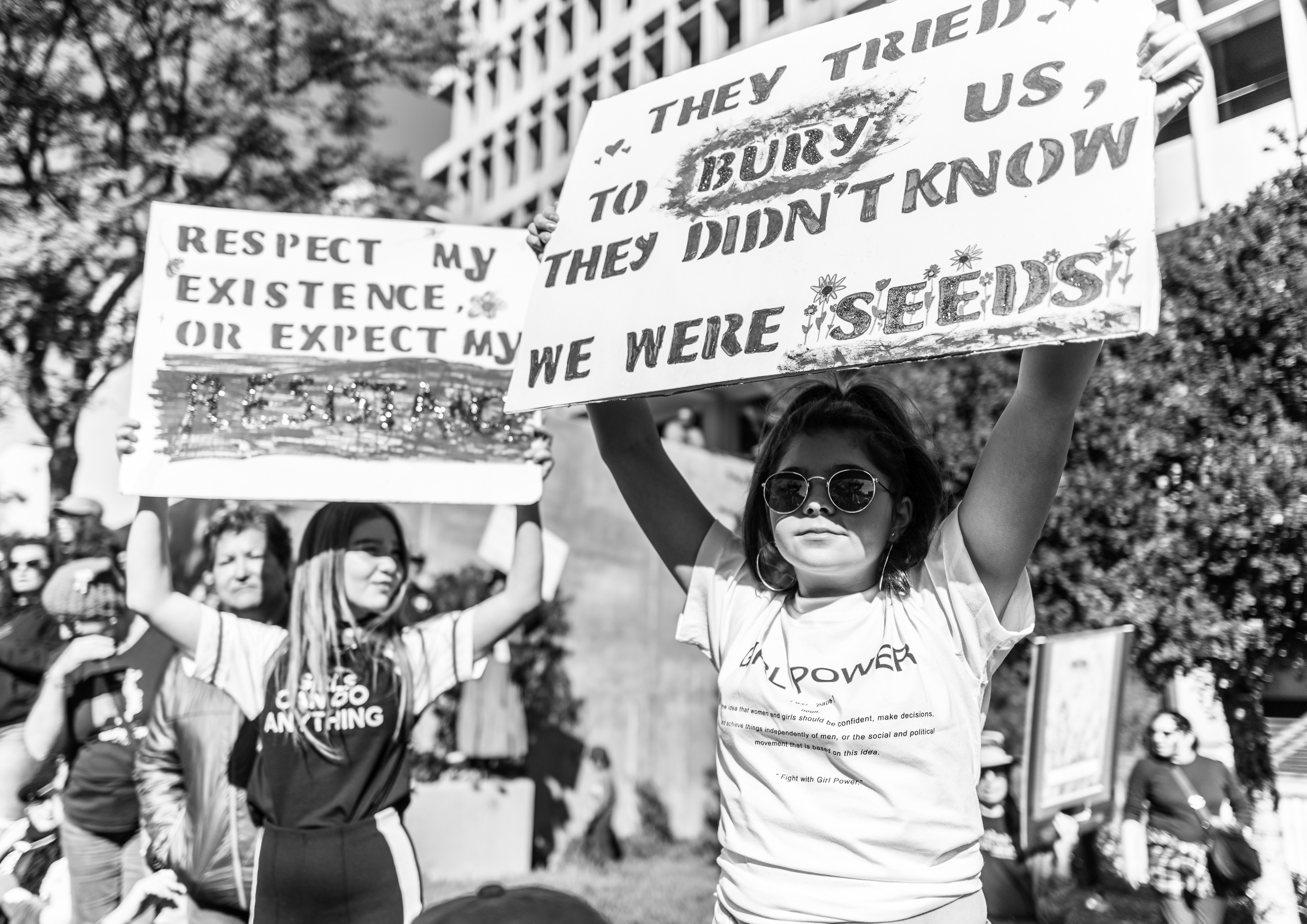 A young girl wearing round sunglasses holds a big sign up in the air. The sign reads, "They tried to bury us, they didn't know we were seeds."