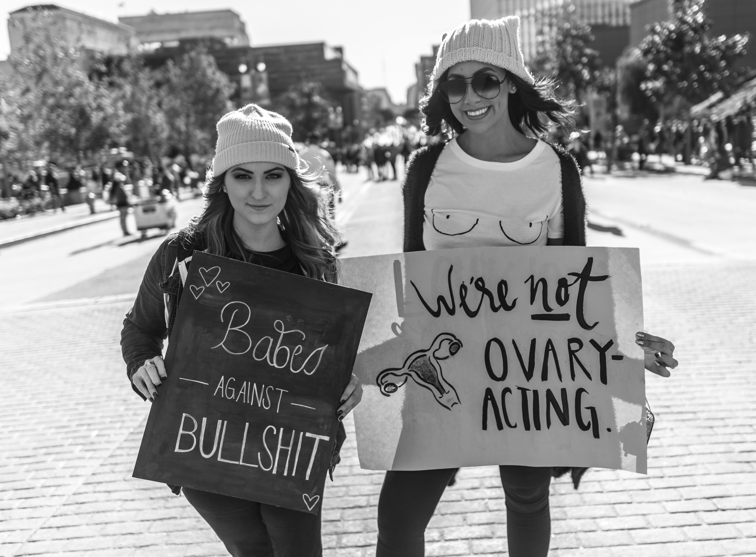 Two friends stand in the street in Downtown Los Angeles at the 2018 Women's March. One holds a sign that reads "Babes against bullshit" and the other holds a sign that reads "We're not ovary acting"
