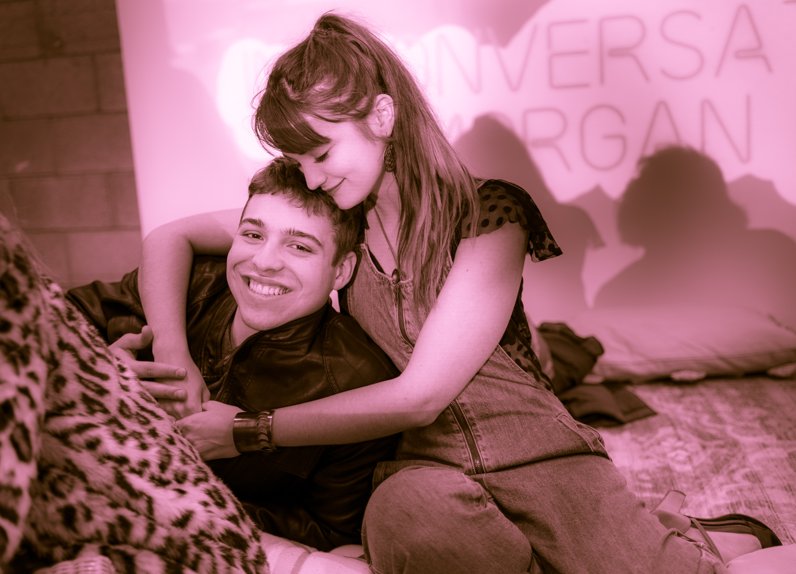 A couple embrace. A guy lays on a pillow, and a girl puts her arms around him and lays her cheek on his head.