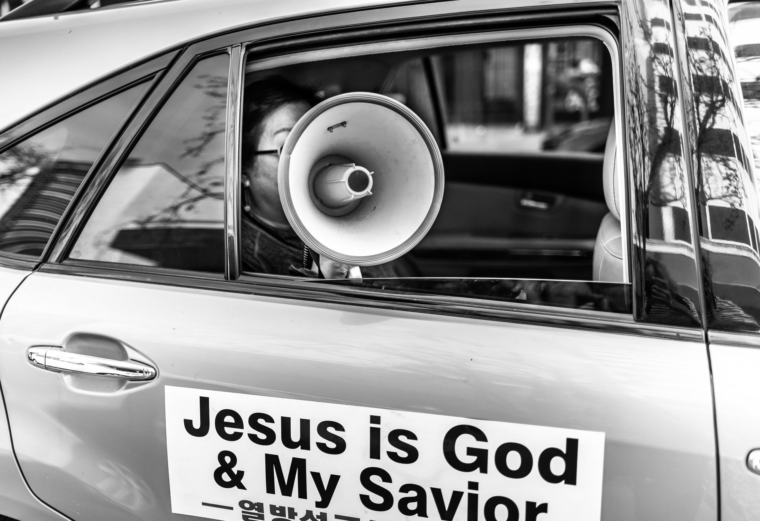 a car drives down a street in Little Tokyo in Los Angeles. A sign on the side of the car reads "Jesus is God & My Savior" in English, and then below that is Korean text, presumably also saying "Jesus is God & My Savior". The rear passenger window is rolled down and a woman holds a megaphone out the window. She speaks various proselytizing text as the car goes down the street. 