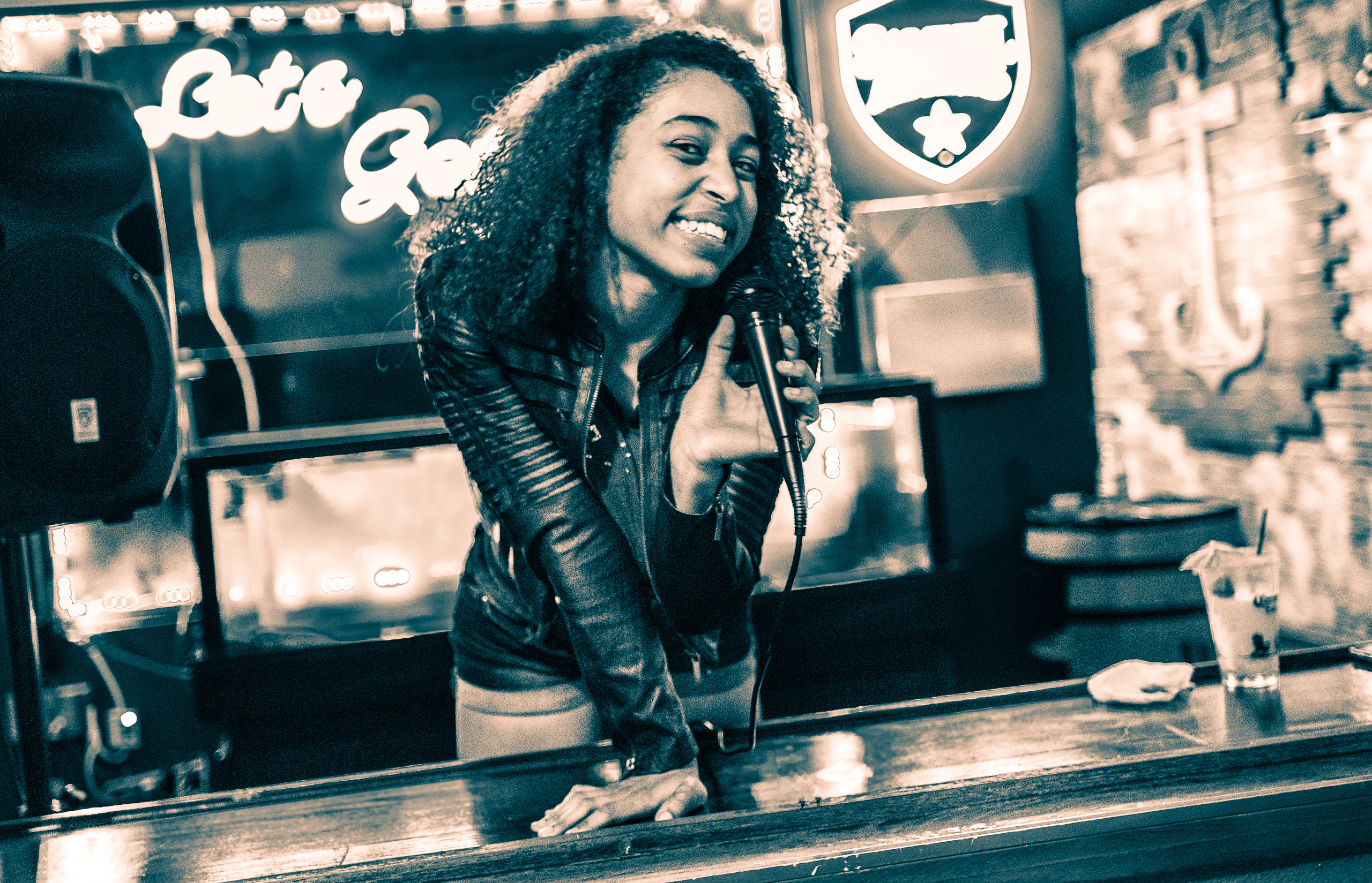 an attractive, young, black woman smiles and talks into a microphone at a streetside window at Tipsy Tails, a bar on Hollywood Blvd. Behind her, a neon sign reads, "Let's Get Tipsy"