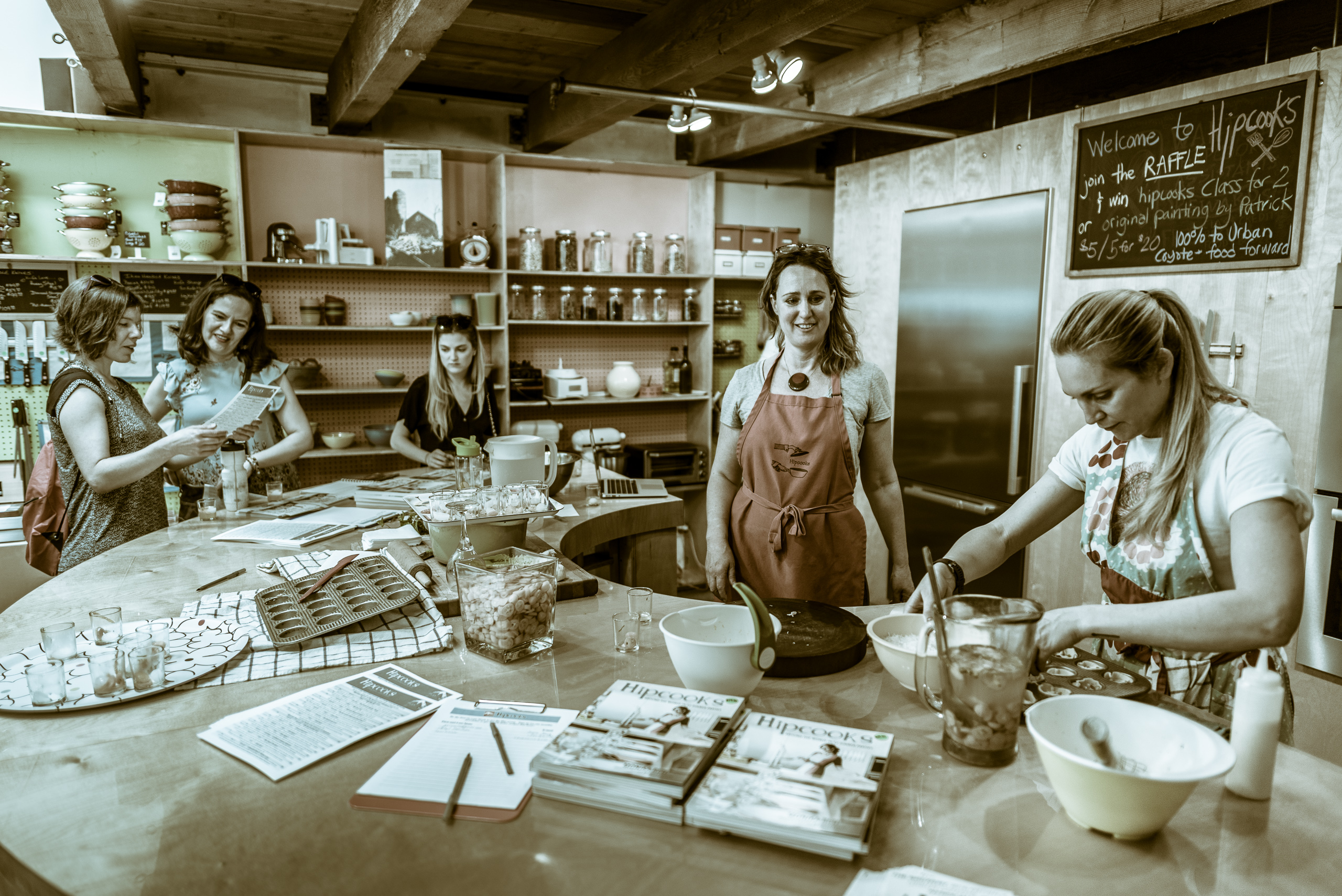 inside the Hipcooks space at The Brewery Artist's Lofts in Downtown Los Angeles. Around a large, semicircular table, one person prepares a dish, one supervises, and another group of 3 examine some items