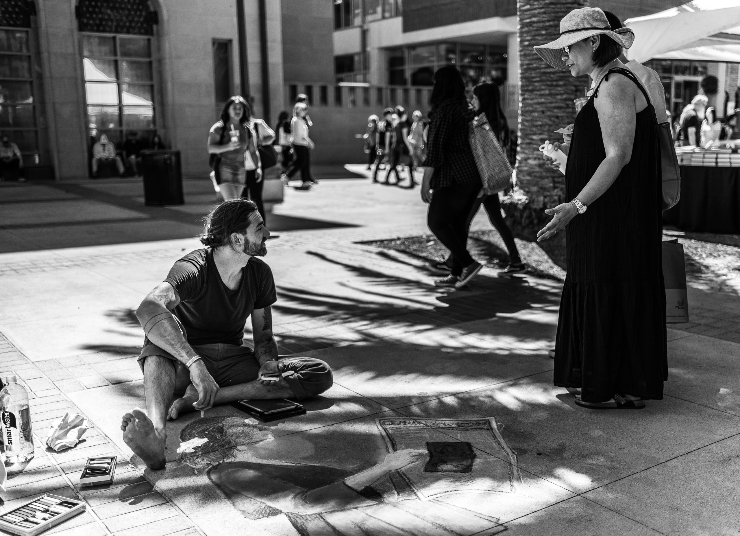 a chalk artist sits on the ground at the University of Southern California campus and works on a chalk/pastel illustration of a young girl in a dress reading the newspaper. A woman gestures toward the artist and tells him how amazing his work is.