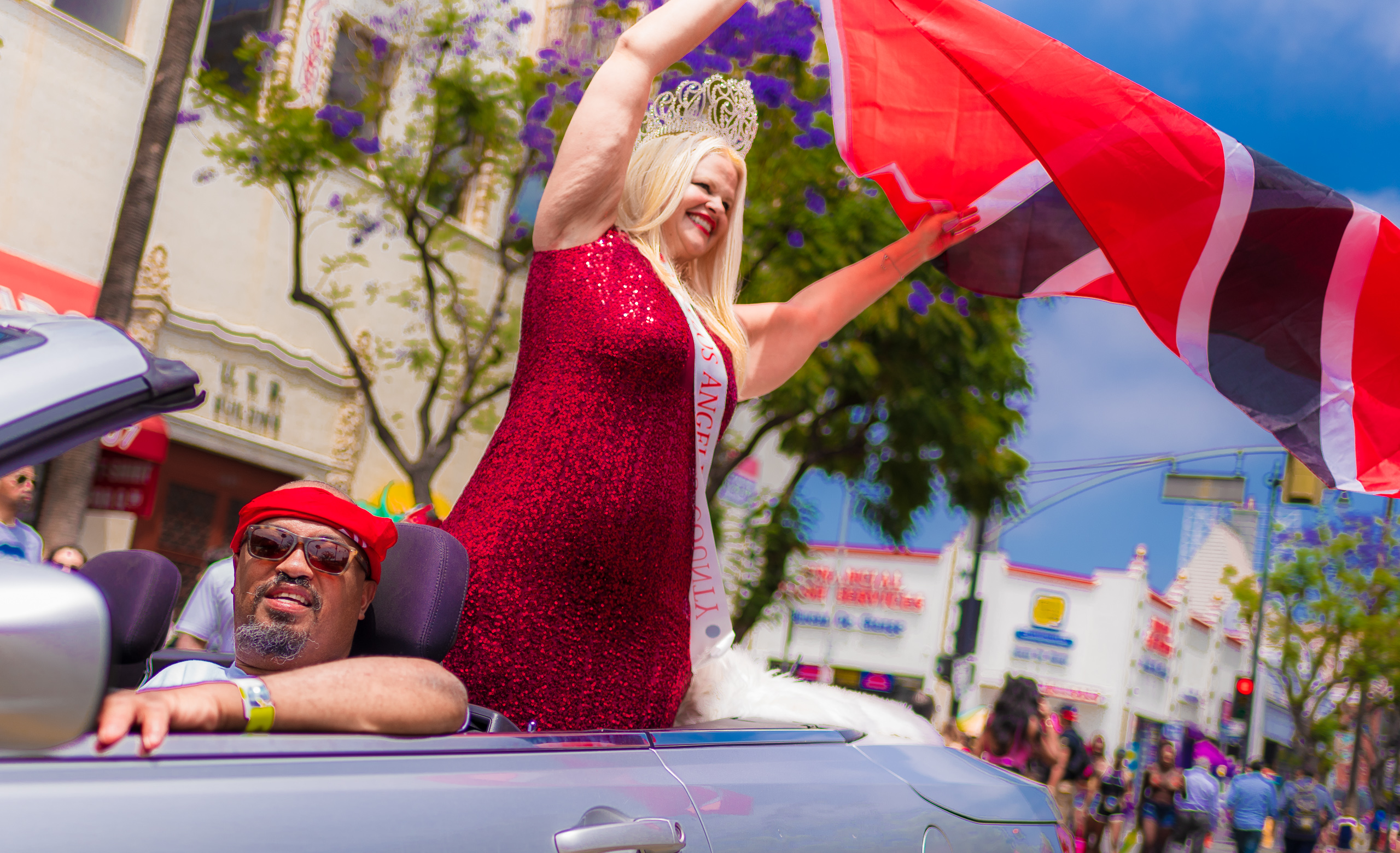 a woman with a bright, sequined red dress and a "miss Los Angeles County" sash waves a Trinidad & Tobago flag as the convertible car she rides in drives down Hollywood Blvd.