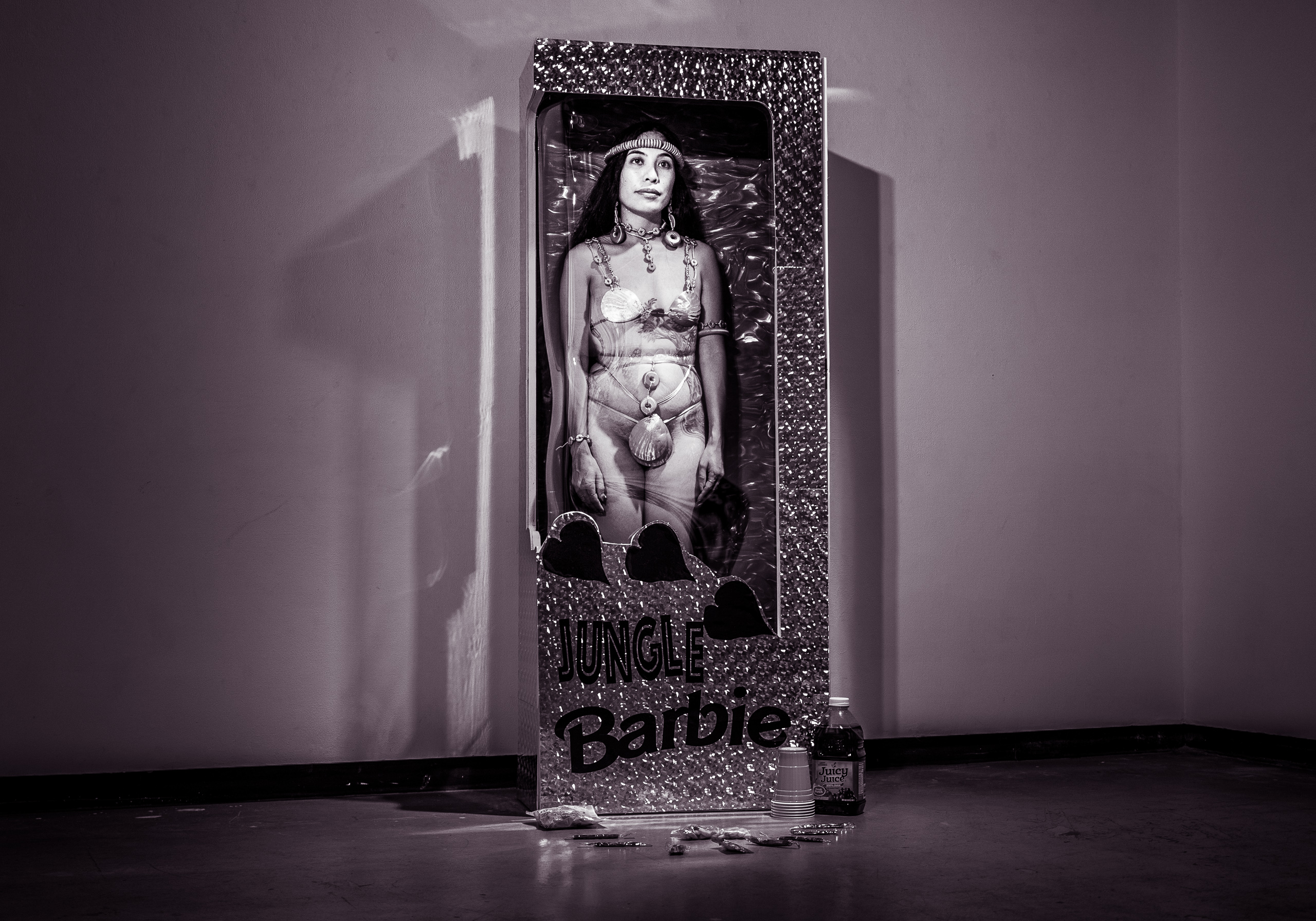 A durational performance by performance artist Lia Gogue performing "Jungle Barbie". Gogue is dressed in a "native" or "jungle" costume and stands inside a life-size "Barbie Box" complete with plastic straps holding the "Barbie Doll" in the box. 