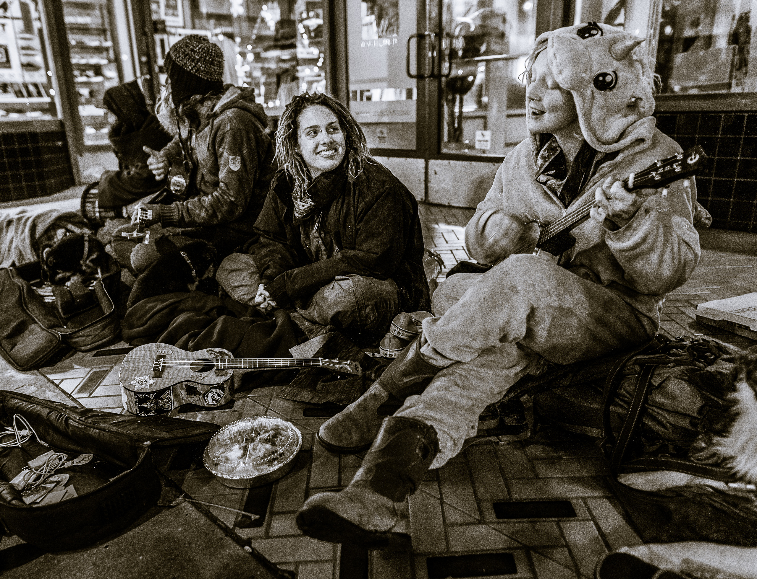 a group of people with food, blankets, guitars, and other materials huddle in the entrance to a storefront on Colorado Blvd in Pasadena on New Year's Eve. Some of them play guitars.