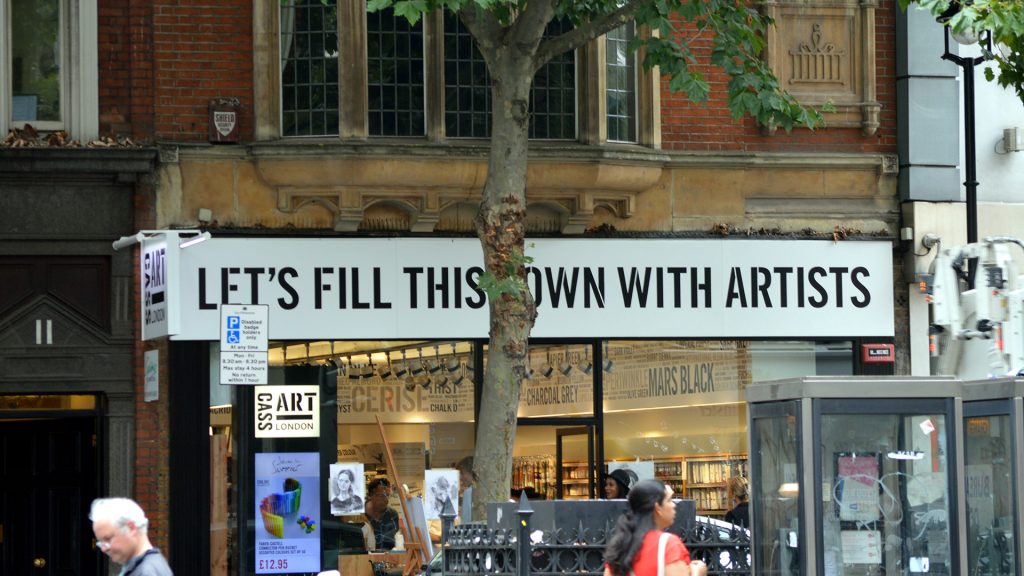 photo of an art store with a large front sign "Let's Fill This Town WIth Artists"