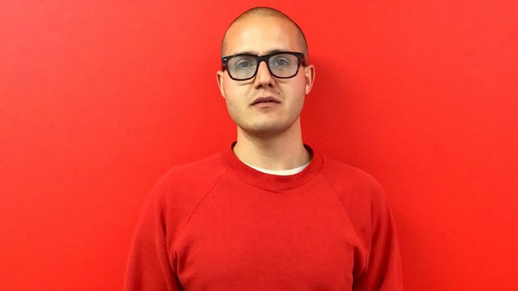 photo of artist Adam Sabolick wearing an orange sweatshirt and standing in front of an orange background